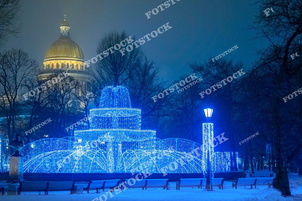 На фотографии изображен Исаакиевский собор на фоне парка, украшенного новогодней иллюминацией. Светящийся фонтан в синем цвете создает сказочную атмосферу зимней ночи. Золотой купол собора эффектно выделяется за деревьями, дополняя композицию величием классической архитектуры. Это место привлекает внимание жителей и туристов в период новогодних праздников.